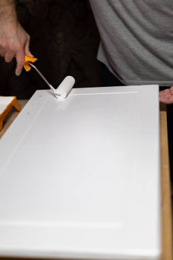 Bir atölyedeki bir dolabın kapısına beyaz boya saran biri DIY ev geliştirme projesini gösteriyor. Kullanılan boya ve aletlerin dikkatli bir şekilde uygulanması üzerinde duruluyor.