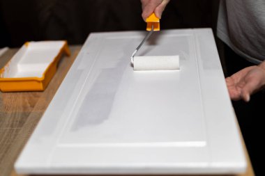 Bir kişi, yeni boyanmış bir boyayı ev dekorasyonu yaparken dolap kapısına sürmek için boya silindiri kullanıyor. Dekor, ahşap yüzeyleri ve minimum dağınıklığı olan rahat bir çalışma alanı..