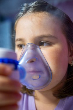 Genç bir kız solunum tedavisi için nebulizör kullanmaya odaklanmış. Cihazı yüzüne yakın tutuyor, ağzını ve burnunu kapatan bir maskeyle..