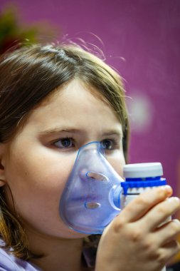 Yüzüne yakın bir nebulizör maskesiyle oturan bir kız. İçerde, sıcak bir ev ortamında sağlık yönetiminin önemli bir anını vurguluyor..