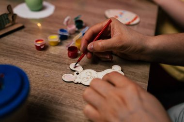 Elleri parlak boyalarla dolu bir masanın üzerine ahşap bir tavşan süsü çizmekle meşgul. Atmosfer rahat ve sanatsal, yaratıcılığı ve ifadeyi teşvik ediyor..