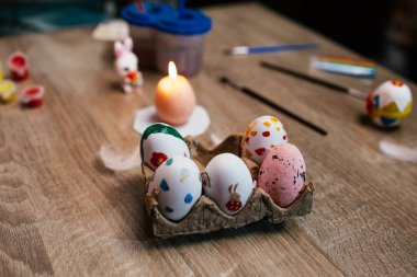 Çeşitli renklerde dekore edilmiş yumurtalar ahşap bir masanın üzerindeki yumurta kutusunda dinleniyor. Yakınlarda bir mum hafifçe yanıp sönüyor ve sanat eserleri bahar kutlamalarıyla ilgili yaratıcı aktivitelere dair ipuçları veriyor..