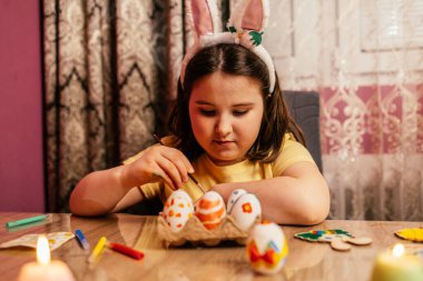 Tavşan kulaklı bir kız evdeki ahşap masaya canlı yumurtalar çizer. Yumuşak mum ışığı, neşeli bahar atmosferine sıcaklık katarken yaratıcı aktivitelerin tadını çıkarıyor..