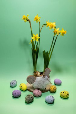 Parlak sarı çiçekler, çeşitli renklerde canlı renkli yumurtalarla çevrili dekoratif bir tencerede dimdik duruyorlar, solgun bir yüzeyde dinleniyorlar, neşeli bir Paskalya atmosferini çağrıştırıyorlar..