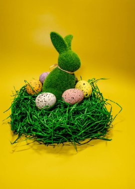 Şenlikli bir sergide yeşil çimenli bir yatakta pastel renkli yumurtaların arasına yerleştirilmiş yeşil bir tavşan sergileniyor. Parlak sarı zemin, Paskalya kutlamaları için mükemmel neşeli bir ortam yaratıyor..