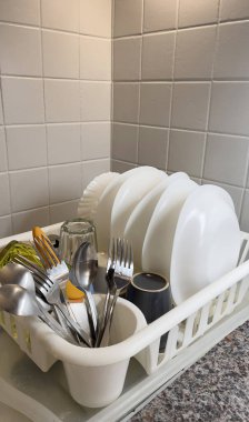 Temiz tabaklar ve çatal bıçaklar, hafif fayanslarla döşenmiş bir duvara karşı kurulmuş kurutma rafında düzgünce düzenlenir. Kurulum, gündüz vakti modern bir mutfak alanında örgütlenmeyi ön plana çıkarıyor.