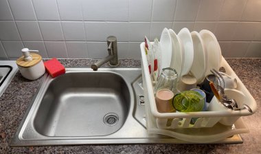 Sıcak bir mutfakta, lavabo temiz bulaşık ve çatal bıçaklarla dolu, canlı renkler sergilenen ve bir sabun makinesinin yanına yerleştirilmiş kuru bir raf tutar..