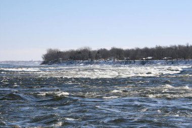 LaSalle, Quebec 'teki Saint Lawrence Nehri' ndeki hızlı kış akıntılarının nefes kesici manzarası. Karla kaplı kıyılar, çıplak ağaçlar ve berrak mavi gökyüzünün altında uzak bir kilise kulesi.