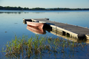 Vaudreuil, Quebec - Durgun suyu yansıtan kırmızı bir tekneyle ahşap bir rıhtımın yer aldığı huzurlu göl manzarası. Etrafındaki sazlıklar ve uzak ağaçlar berrak gökyüzünün altındaki sakin atmosfere katkı sağlar.
