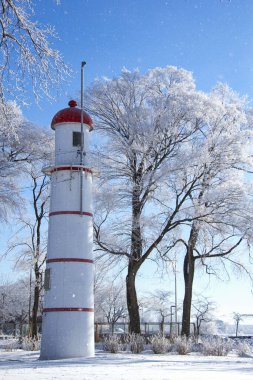 Scenic winter view of a white and red lighthouse surrounded by frosted trees under a clear blue sky, with snowflakes gently falling clipart