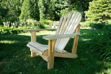 Çok güzel işlenmiş ahşap bir Adirondack sandalyesi yeşil bir çimenlikte, yemyeşil bitkilerle ve huzurlu bir bahçe ortamında çiçek açan çiçeklerle çevrili. Rahatlamak ve doğanın tadını çıkarmak için mükemmel bir yer.
