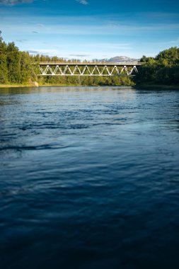 Kuzey Norveç 'te, parlak mavi gökyüzünün altında, karanlık bir nehir boyunca çelik bir köprü, ön plandaki nehir.