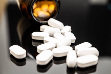 Koyu kehribar reçeteli bir şişeden yansıtıcı bir yüzeye dökülen beyaz afyon haplarının yakın plan görüntüsü, fentanil gibi aşırı dozda opioid ve reçeteli ilaçların zararlarını vurguluyor..