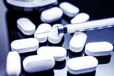 İlaçlar, sağlık hizmetleri ve tıbbi tedavi temalarını vurgulayan, yansıtıcı karanlık bir yüzeyde sıvı bulunan bir şırınganın yanına saçılmış beyaz hapların detaylı bir yakın çekimi..