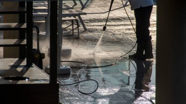 Outdoor cleaning scene captures a worker using a high-pressure water jet to clean a sunlit patio, highlighting water reflections and safety gear among architectural elements. clipart