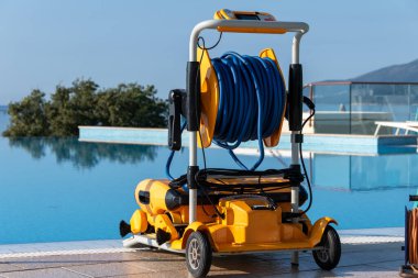 Uzun mavi sarmal hortumlarla donatılmış sarı bir robotik havuz temizleyicisi sakin, berrak dağlara bakan mavi bir havuzun yanına park etmiş. Gelişmiş havuz bakımı ve temizlik ekipmanları lüks bir açık havada sergilendi..