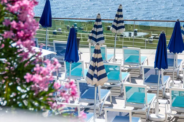 stock image An inviting seaside resort terrace featuring rows of turquoise sunbeds matched with stylish blue and white striped umbrellas. Elegant relaxation by the clear blue ocean, framed by vibrant pink flowers.