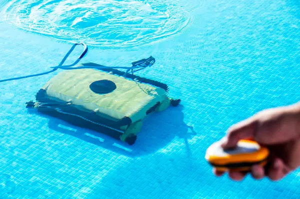 stock image A robotic pool cleaner works efficiently in a clear blue swimming pool, operated via remote control by a person's hand, emphasizing convenience and modern pool maintenance technology.