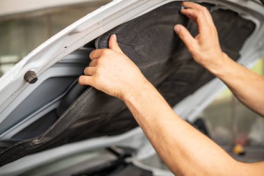 Bir tamircinin elleri dikkatle bir aracın motor bölmesindeki alt kaput yalıtımını kaldırarak ve değiştirerek, gürültüyü azaltarak ve bileşenleri ısıdan koruyarak optimal performansı sağlar. Otomotiv tamiri ve bakım temaları için ideal.