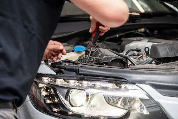 stock image A skilled technician is diligently inspecting the engine compartment of a modern vehicle. Using a screwdriver, the mechanic works on crucial components, ensuring optimal performance for the car. Garage setting highlights automotive expertise.