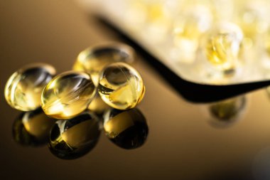 D vitamini ve Omega-3 balık yağı içeren sarı şeffaf kapsüllerin yakın görüntüsü, sağlık ve refah kavramları için ideal yansıtıcı bir yüzeyde sergilenmektedir..