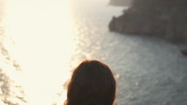 Arkadan yakından bakınca denize ve yukarıdan kayalık kıyılara bakan bir kız görülüyor. Saçları rüzgarda savrulurken nefes kesen manzarayı alıyor..