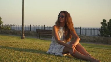Güzel bir kız yeşil çimenlerde oturuyor, günbatımının tadını çıkarıyor. Batan güneşin sıcak, altın ışığı onu yumuşak bir ışıltıyla yıkar, o anın dinginliğini ve memnuniyetini arttırır. 4k görüntü