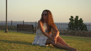Güzel bir kız yeşil çimenlerde oturuyor, günbatımının tadını çıkarıyor. Batan güneşin sıcak, altın ışığı onu yumuşak bir ışıltıyla yıkar, o anın dinginliğini ve memnuniyetini arttırır. 4k görüntü