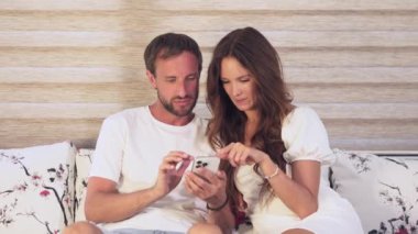 Genç Avrupalı çift kanepede oturup evde birlikte alışveriş yaparken akıllı telefon cihazları kullanarak eğleniyor. Gülümseyen genç karı ve koca gülüyor. Cep telefonu cihazları üzerinde evde gezinme uygulamasında rahatlayın.. 