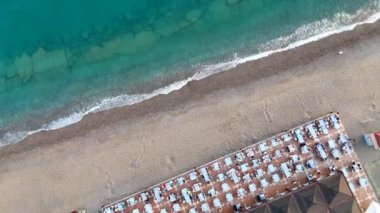 İnsansız hava aracı perspektifi, gün batımının ılık renkleriyle yıkanmış sakin bir Akdeniz plajını gösteriyor. Kıyıya vuran yumuşak dalgaları ve sudan yansıyan canlı renkleri vurguluyor.