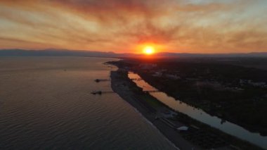 Sersemletici turuncu günbatımının altında bir nehrin denizle birleştiği sakin bir manzara. Etrafındaki dağlar, suların yüzeyindeki sıcak ışığı yansıtan güzelliği arttırır.
