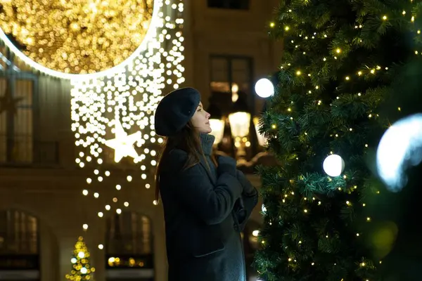 Gri paltolu ve klasik bereli Parisli bir kadın, Noel ağacındaki yeni yıl oyuncaklarına hayranlık duyuyor ve Paris 'teki tatil sezonunun neşeli atmosferinin tadını çıkarıyor.