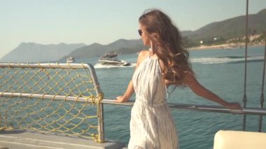 Kız bir yatın güvertesinde, denizde sorunsuzca süzülüyor. Arka planda görkemli dağlar yükselirken, uzaklardan bir tekne geçiyor. Güneş sulardan parıldıyor, dinginlik katıyor.