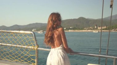 Kız, sakin sularda süzülen bir yatta zarifçe duruyor. Arka planda görkemli dağlar yükselirken, küçük bir tekne geçiyor. Sahne, dinginlik ve doğal güzelliği yakalar.