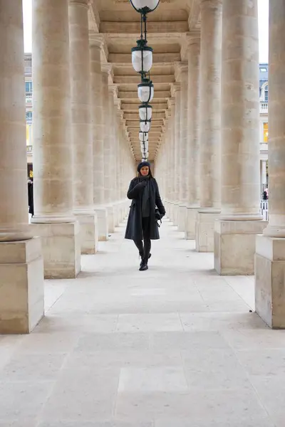 Paris 'teki Palais Royal' in büyük sütunlu geçidinde şık bir kış kıyafeti giymiş bir kadın yürüyor, Paris 'in zarafetini ve tarzını tarihi bir ortamda somutlaştırıyor. Yüksek kalite fotoğraf