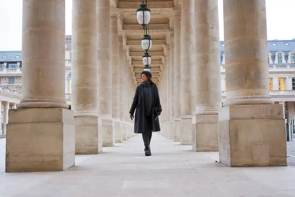 Kış kıyafetleri içinde şık bir kadın Palais Royal 'in taş sütunları arasında yürüyor. Simetrik tasarım ve Paris tarzı, zamansız, zarif bir sahne yaratıyor.