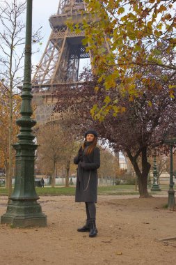 Bere ve palto giymiş bir kadın Eyfel Kulesi yakınlarındaki renkli sonbahar ağaçlarının altında parkta geziniyor. Onun şık Paris tarzı çevrenin zarafetine uyuyor..