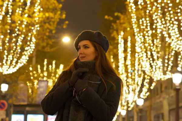 Bere ve eldiven giymiş sıcacık giyinmiş bir kadın Paris sokaklarında şenlikli tatil ışıklarının arasında duruyor. Parlayan ışıklar ve rahat elbisesi sıcak, büyülü bir gece atmosferi yaratıyor. Kaliteli fotoğraf