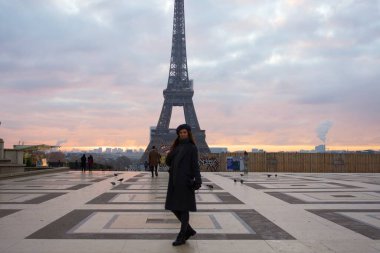 Dressed in chic winter attire, a woman poses against the backdrop of the Eiffel Tower during a peaceful Parisian sunrise. High quality photo clipart