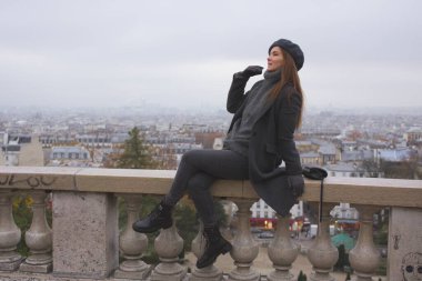 Paris 'e bakan bir kadın Montmartre' daki balyalara yaslanıyor. Kış kıyafetleri ve beresi aşağıdaki şehrin güzel, puslu manzarasını tamamlıyor..