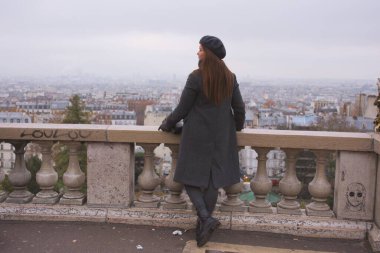 Kışlık paltolu ve bereli bir kadın Montmartre 'dan Paris' in geniş siluetine bakıyor. Bulutlu gökyüzü bu pitoresk şehir manzarasına huzur ve yansıtıcı bir hava katıyor..