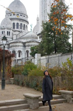 Montmartre, Paris 'teki Sacre-Cour Bazilikası önünde rahat kışlık ceketli neşeli bir kadın poz veriyor. Sonbahar yaprakları ve yeşilliklerle çevrili bu ikonik zemin karşısında mutluluk saçıyor.