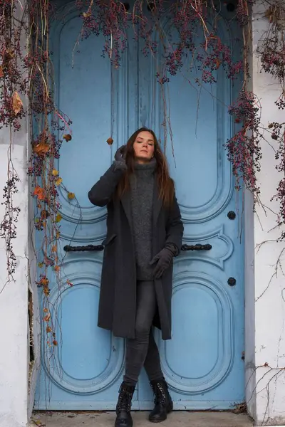 Gri ceketli ve eldivenli bir kadın, Montmartre, Paris 'te sarmaşık tarafından çerçevelenmiş parlak mavi bir kapının yanında duruyor. Sahne, mevsimlik cazibeyle Paris zarafetini birleştirir.