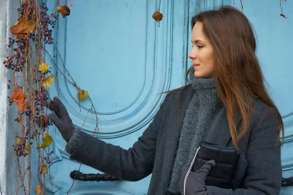 Sıcak kış kıyafetleri içinde, bir kadın Montmartre, Paris 'te şelaleden sarmaşıklarla dolu canlı mavi bir kapıya karşı güven içinde poz verir ve taşralı Paris cazibesiyle dolu şık bir sahne yaratır.