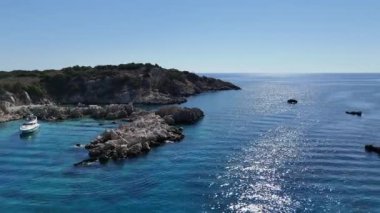 Hareketli bir İHA tarafından yakalanan bu görüntüde ıssız bir körfeze demirlemiş ıssız bir turist yatı, sakin sular ve uzak bir adanın doğal güzelliğiyle çevrili. Kaliteli 4k görüntü