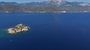 Yukarıdan çekilen bu çarpıcı dron atışı, yüksek dağların dramatik zeminine karşı kurulmuş açık denizle çevrili küçük bir adayı ortaya çıkarıyor. Güneş ışıl ışıl parlarken.