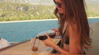 Şık siyah mayo giymiş genç bir kız, bir yat barında eğleniyor, neşeli bir dansla bardaklara kola dolduruyor. Parlak, güneşli bir günün ve ışıl ışıl okyanusun arka planında.