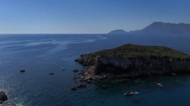 Uçsuz bucaksız mavi denizi, ön planda engebeli kayalık bir adayla ve arka planda yükselen dağlarla kaplayan nefes kesici bir dron atışı. Yüksek kalite 4k görüntü