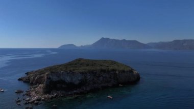 Geniş bir insansız hava aracı görüntüsü mavi denizin, engebeli bir kayalık adanın ve arka planda yükselen dağların çarpıcı panoramik görüntüsünü ortaya çıkarıyor.. 
