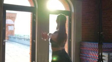 Modern spor giyimli bir balerin, altın saat boyunca güneşli bir çatı katı tarzı spor salonunda zarif bir dans gösterisi yapar. Sıcak günbatımının ışığı büyük endüstriyel pencerelerden geçerken